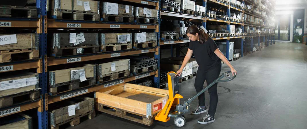 Frau schiebt einem Hubwagen unter eine Palette, um Waren aus einem Regal zu holen